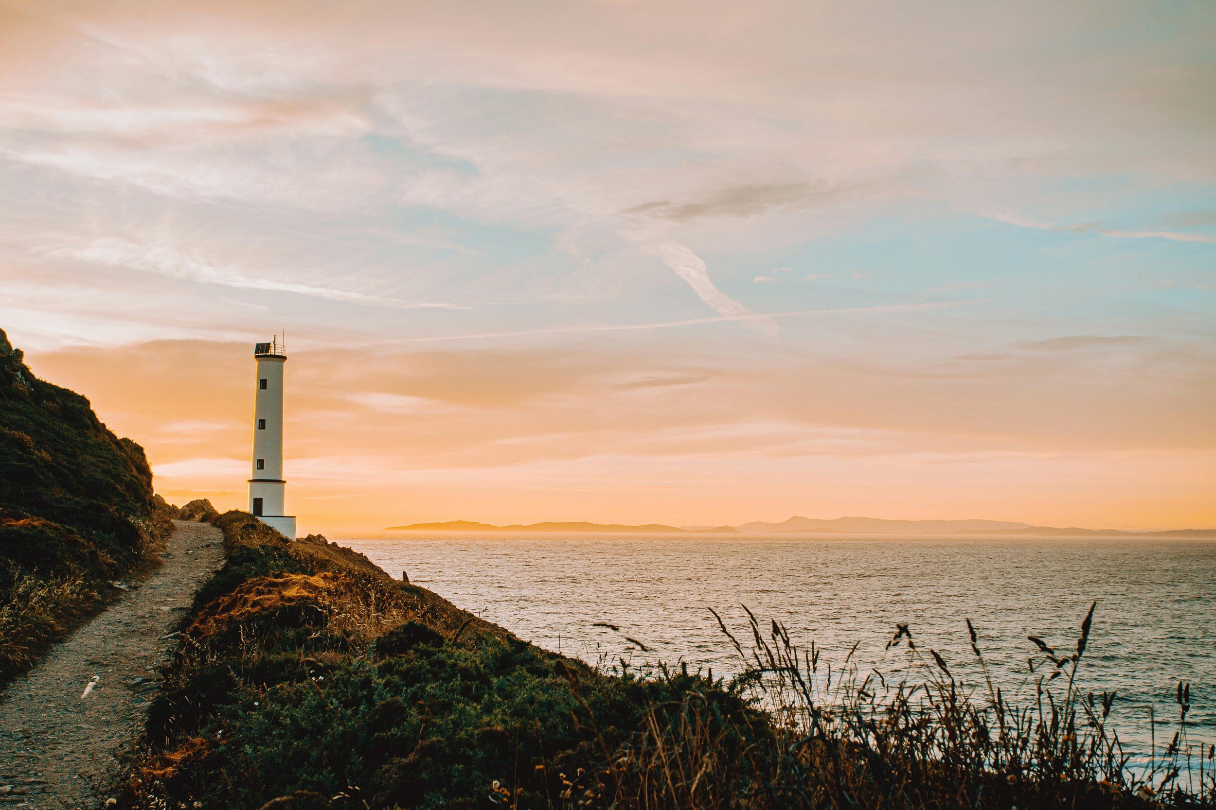 A white lighthouse on a cliff path above the sea at dusk